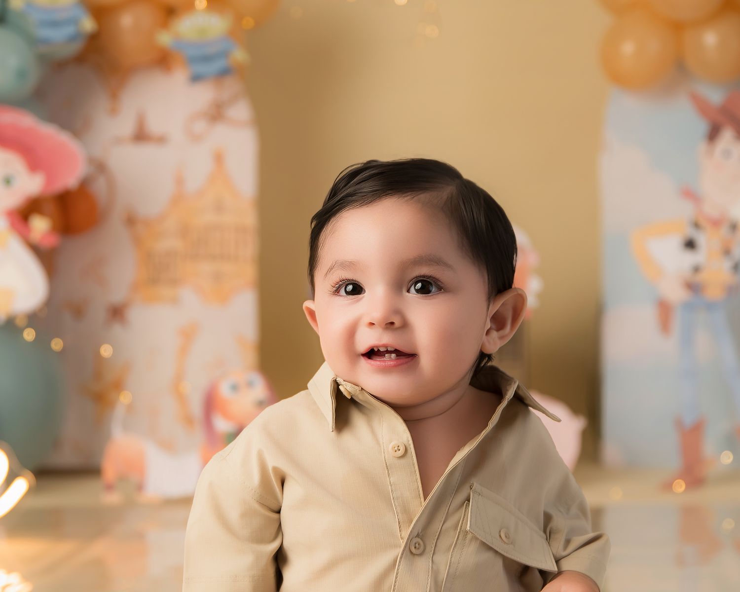 Adorable baby boy in beige outfit smiling at first birthday photoshoot with colorful balloon decorations.