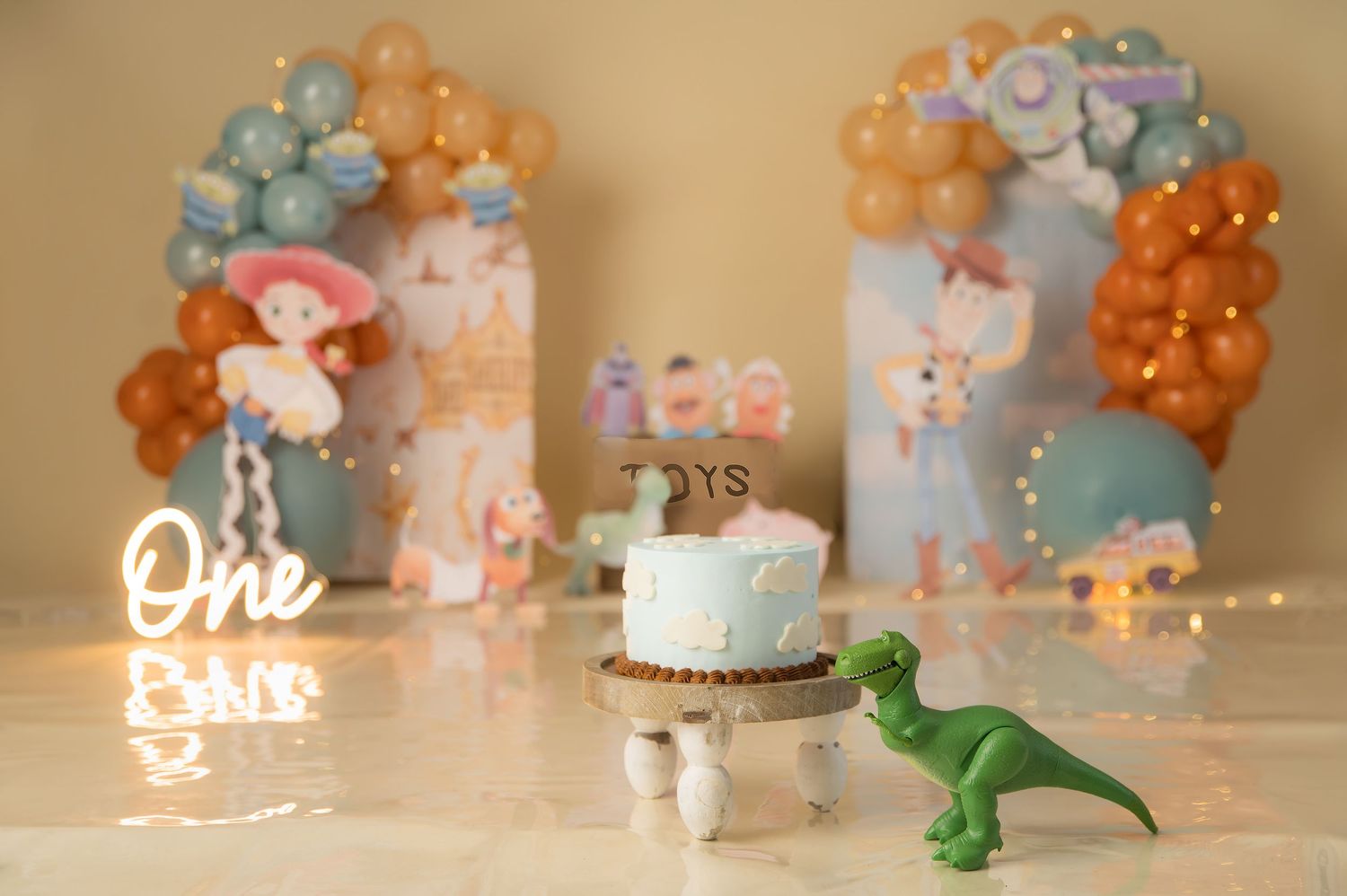 Toy Story themed first birthday cake smash setup with balloon arch, Rex dinosaur toy, and glowing One sign.