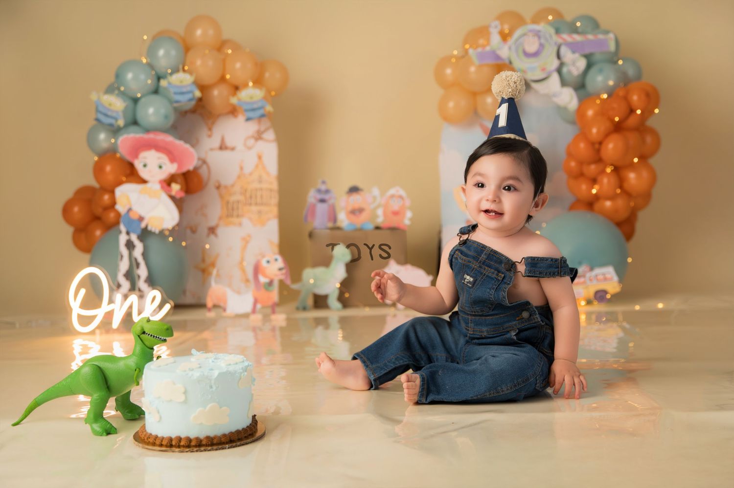 Baby's first birthday cake smash with Toy Story balloons, denim outfit, and party hat on beige backdrop.