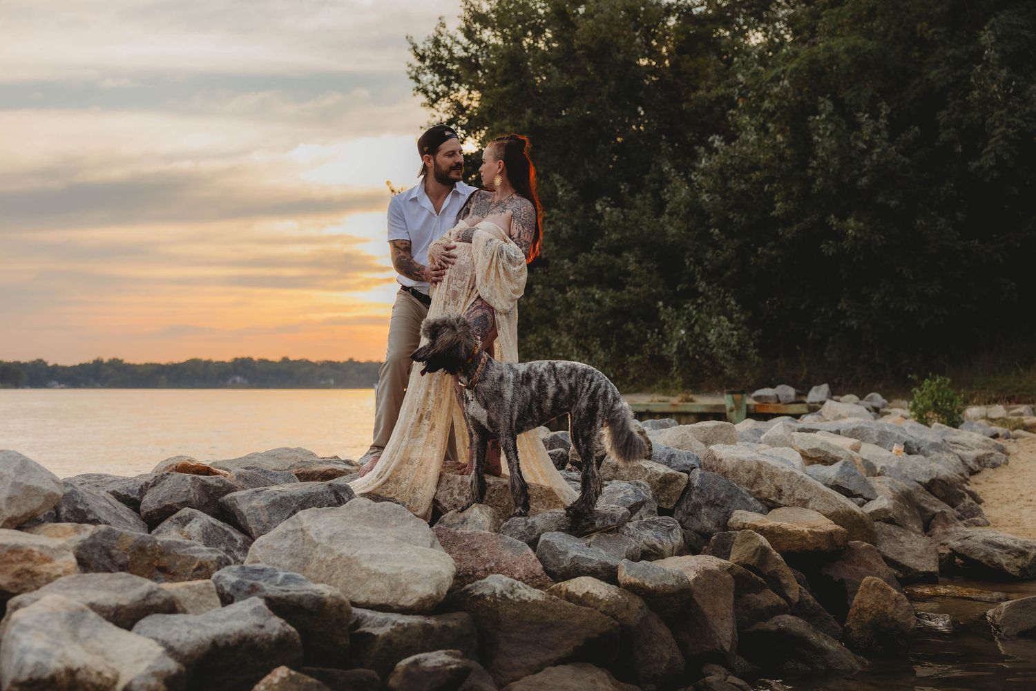 Couple embracing with their Great Dane dog on rocky lakeside shore at golden sunset.