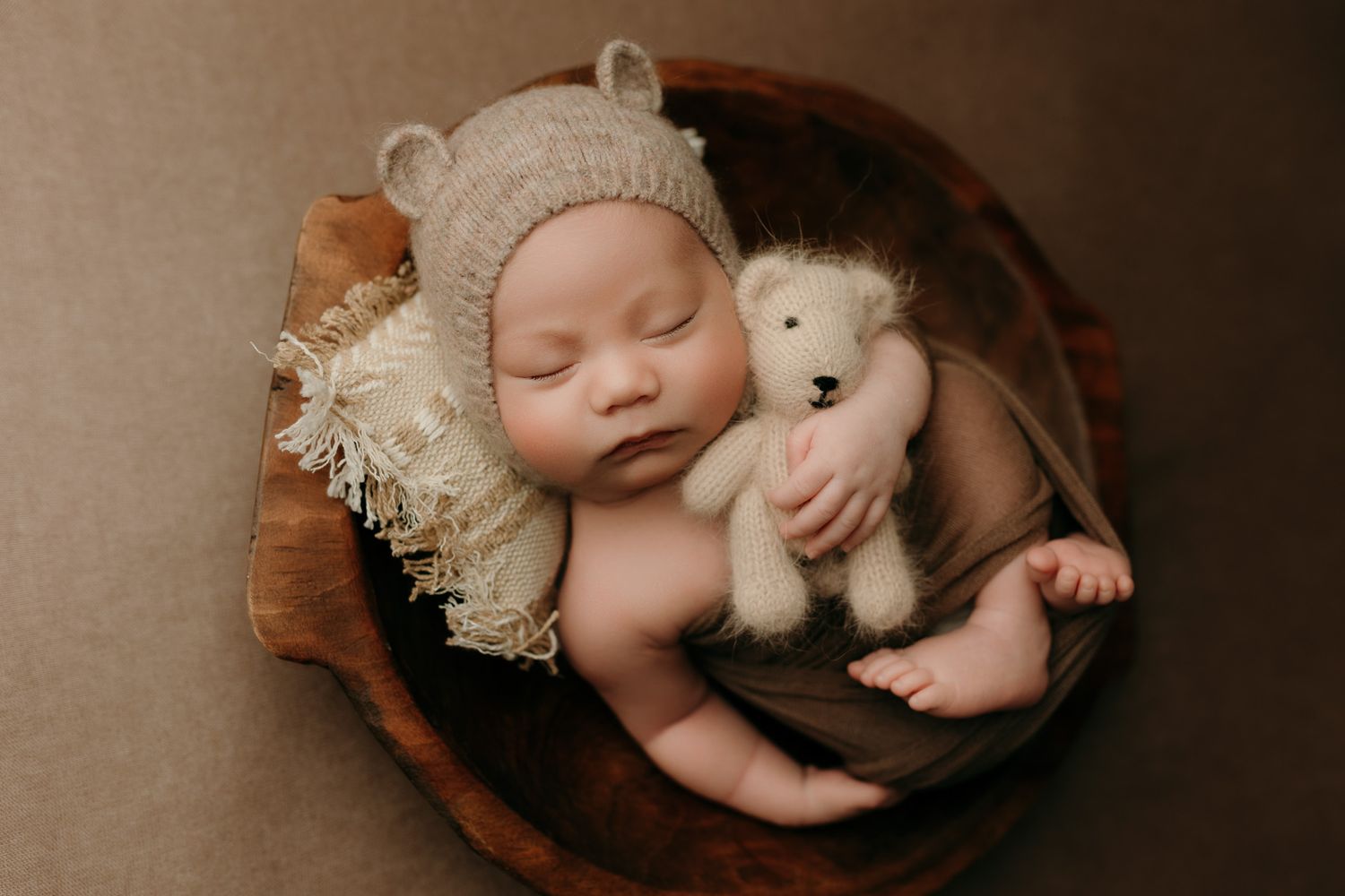 Sleeping baby wearing knit bonnet cuddles with plush bunny toy in wooden bowl during newborn photography session.