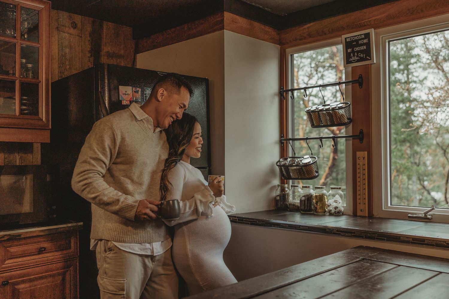 Couple sharing an intimate moment in a rustic kitchen with warm lighting and wooden cabinets near a window.