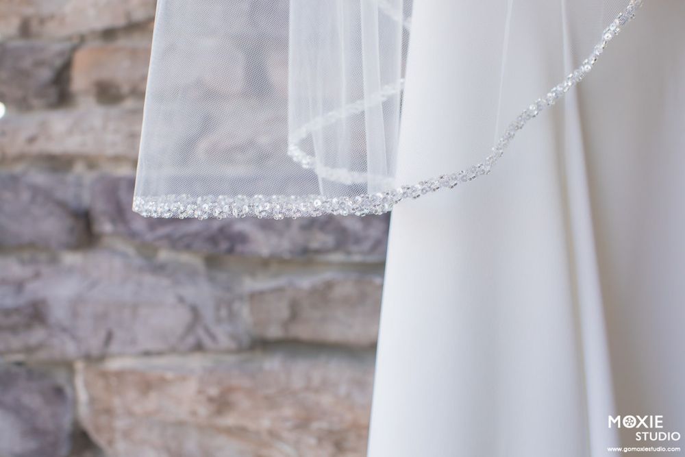 Close-up detail of a delicate white wedding veil with beaded trim against a stone wall background.