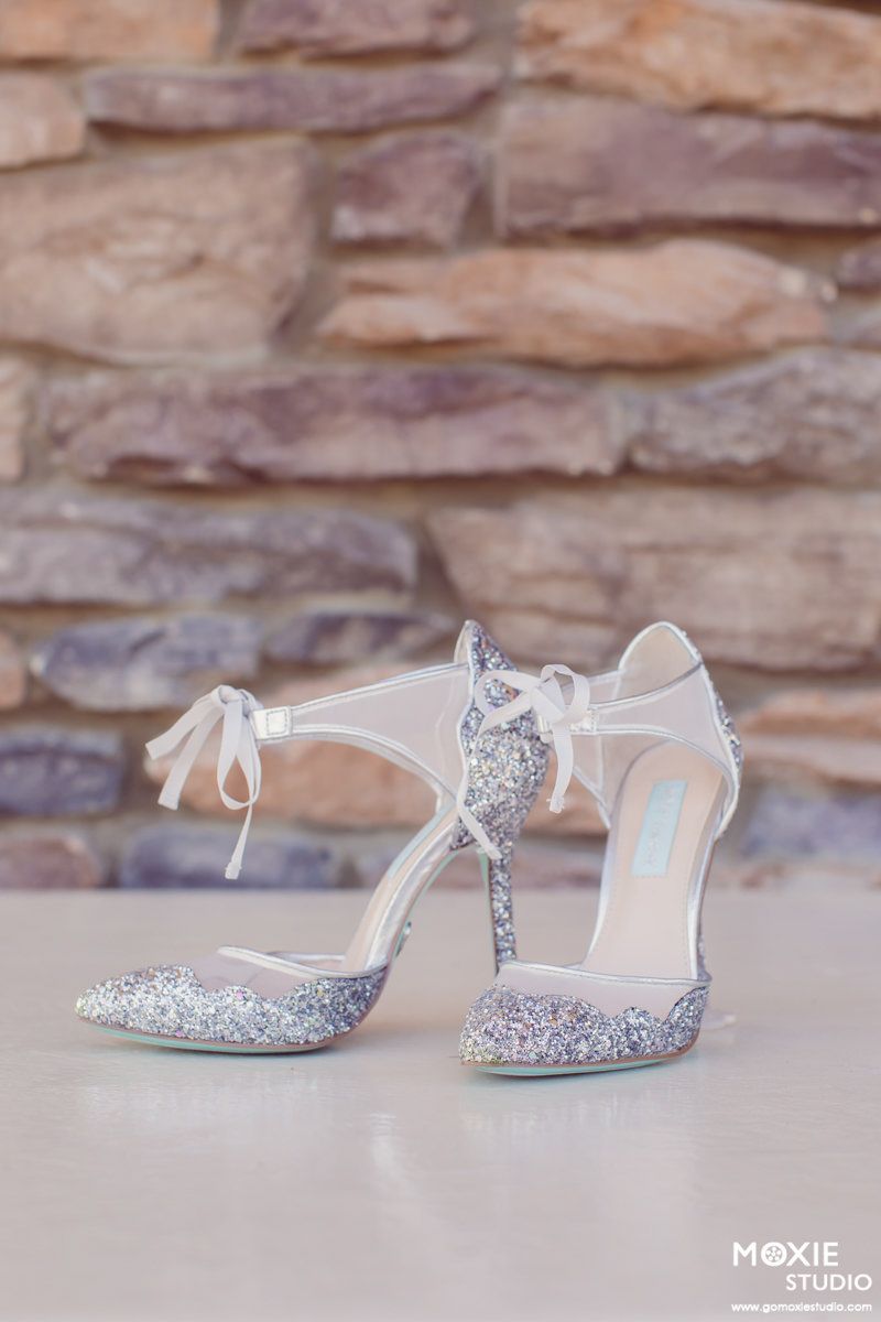 Elegant bridal shoes with silver glitter and white ankle straps sit against a stone wall backdrop.