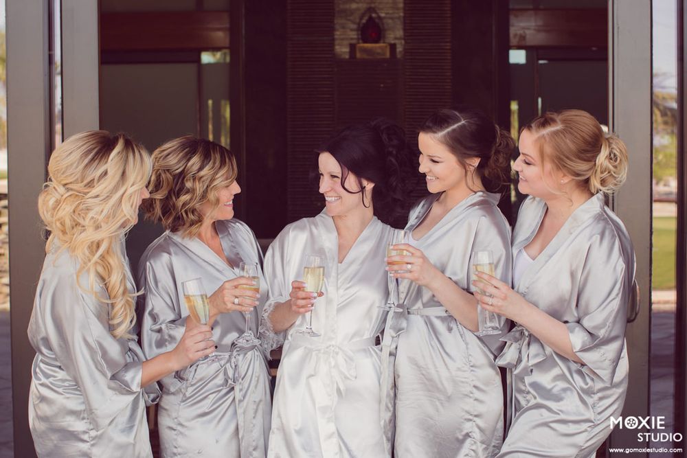 Bridal party in matching silver silk robes laughing together while getting ready for wedding day.