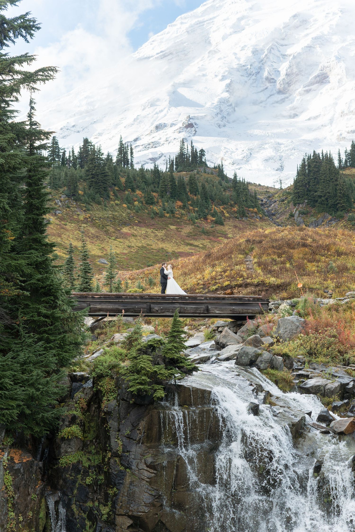 A couple stands on a wooden bridge overlooking a cascading waterfall at Mount Rainier National Park in autumn.