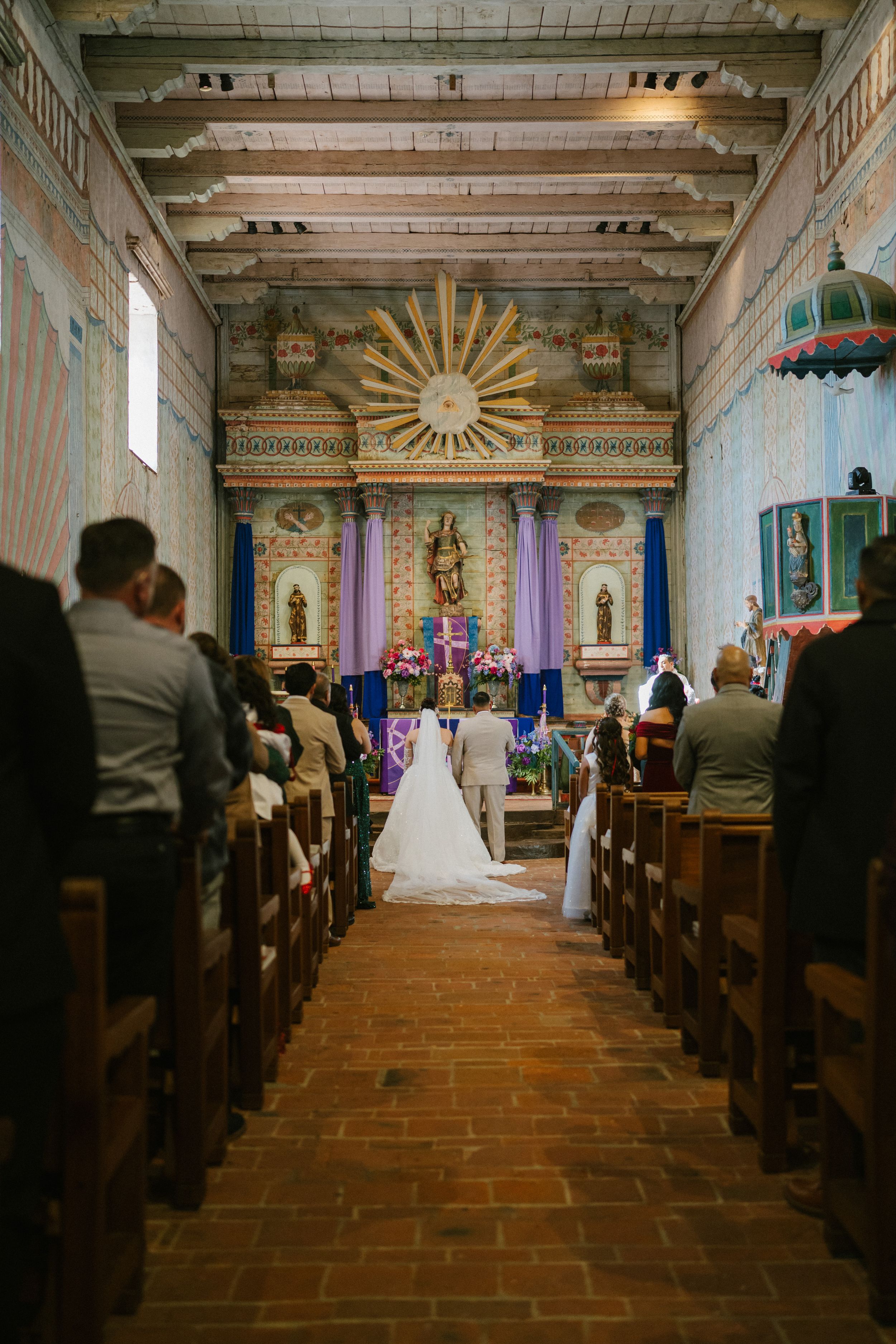 A traditional church wedding ceremony takes place in a historic mission chapel with terra cotta floors and wooden pews.