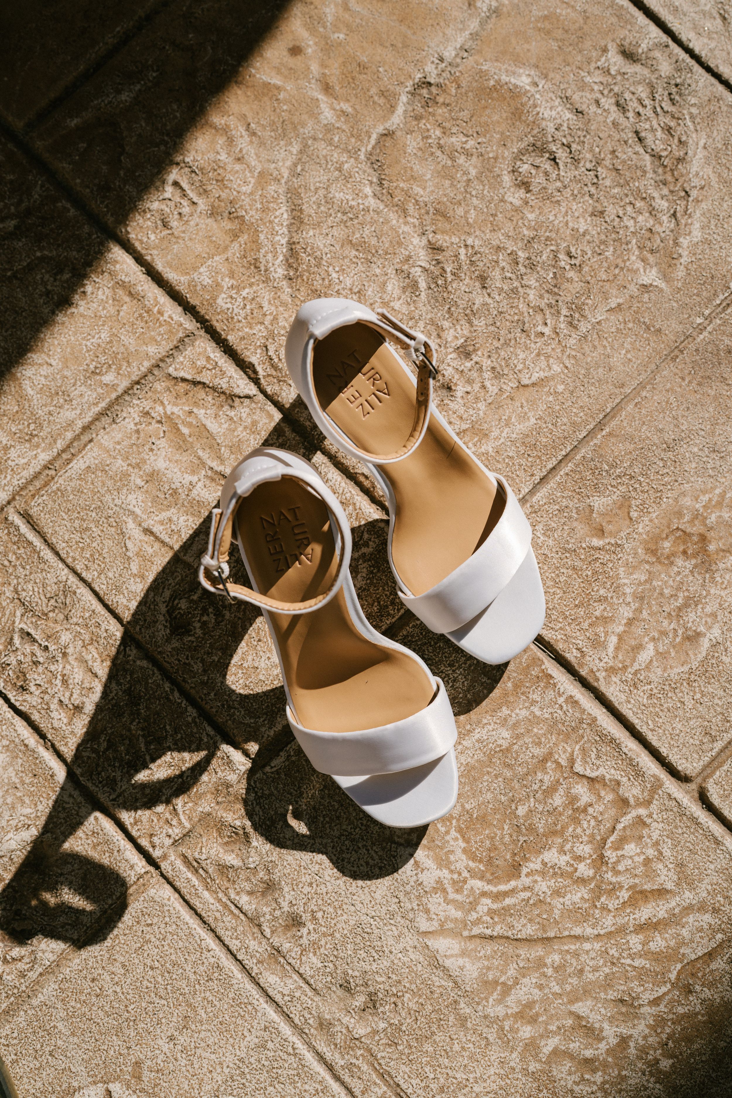 White leather sandals with ankle straps cast dramatic shadows on textured beige stone flooring in sunlight.