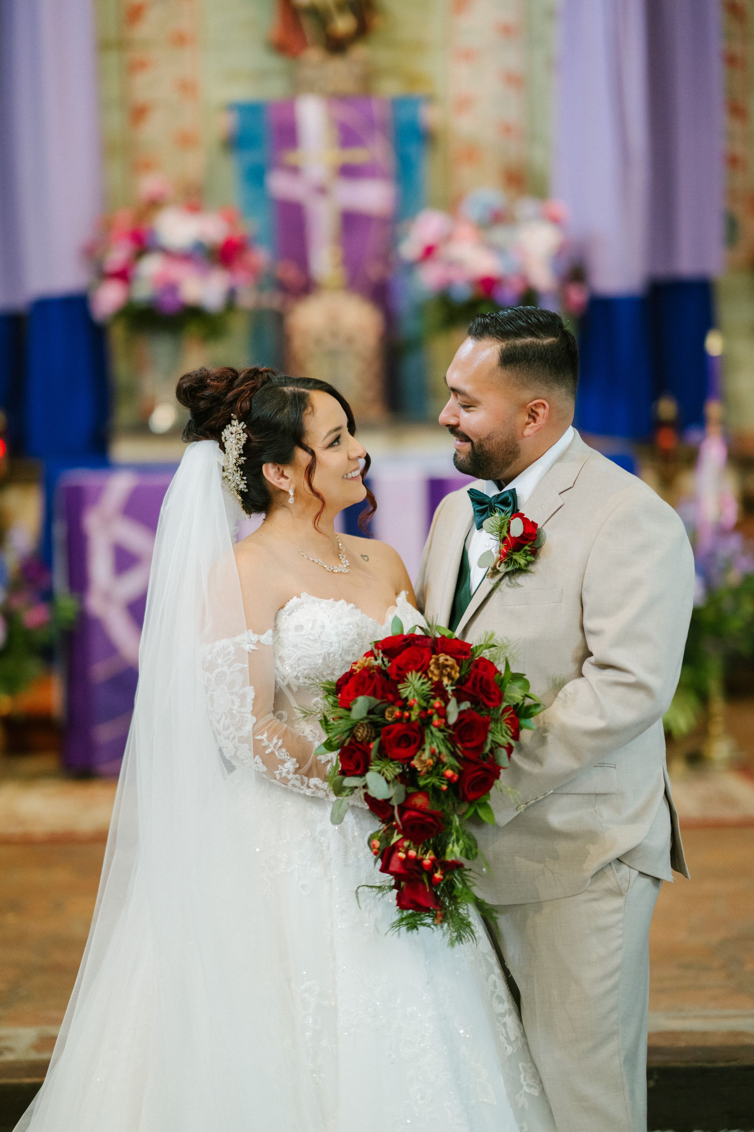 A wedding couple shares a romantic moment in a church with red roses and decorative purple and blue backdrop.