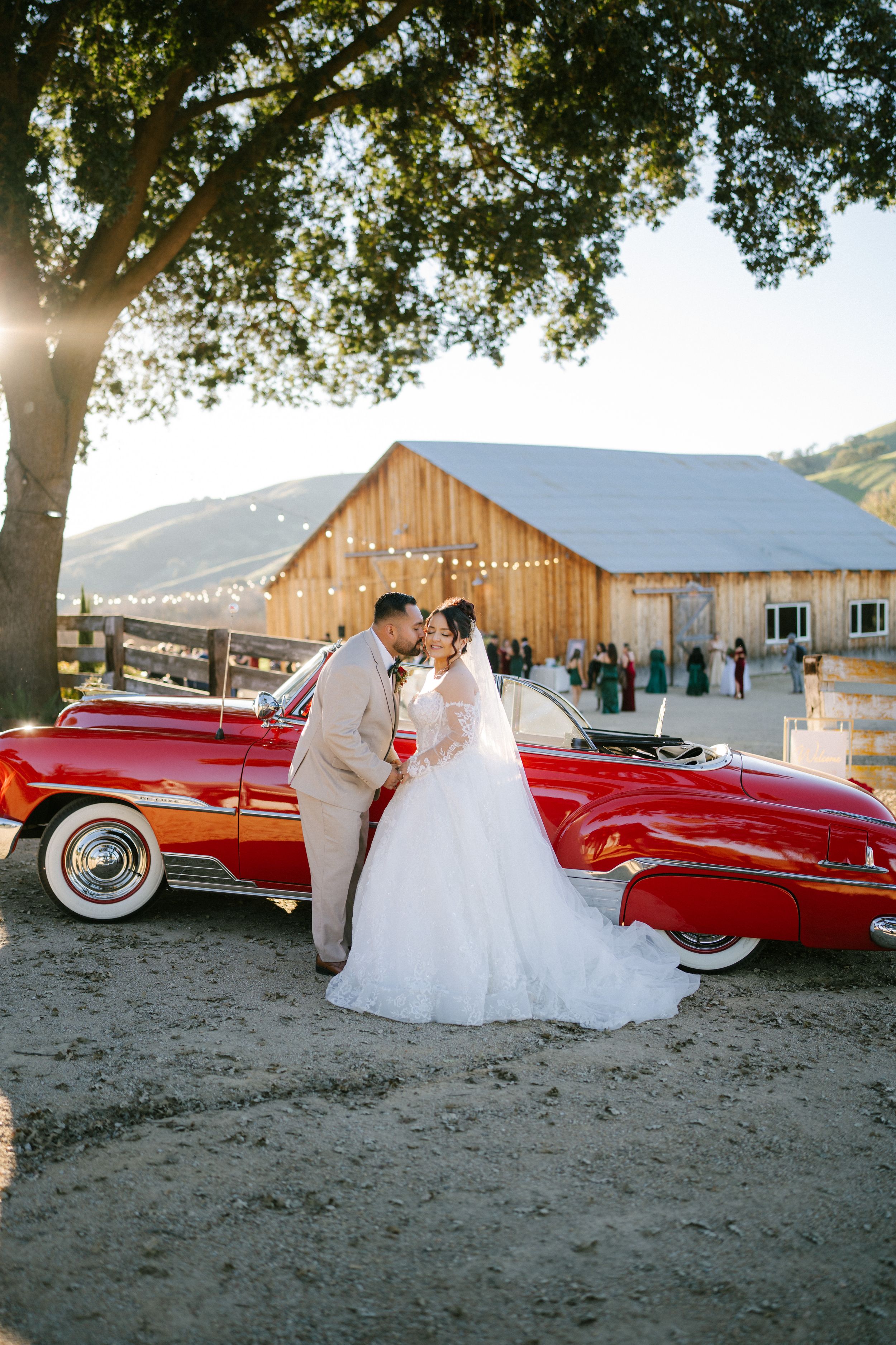 A romantic wedding scene with a vintage red convertible car parked in front of a rustic barn venue.