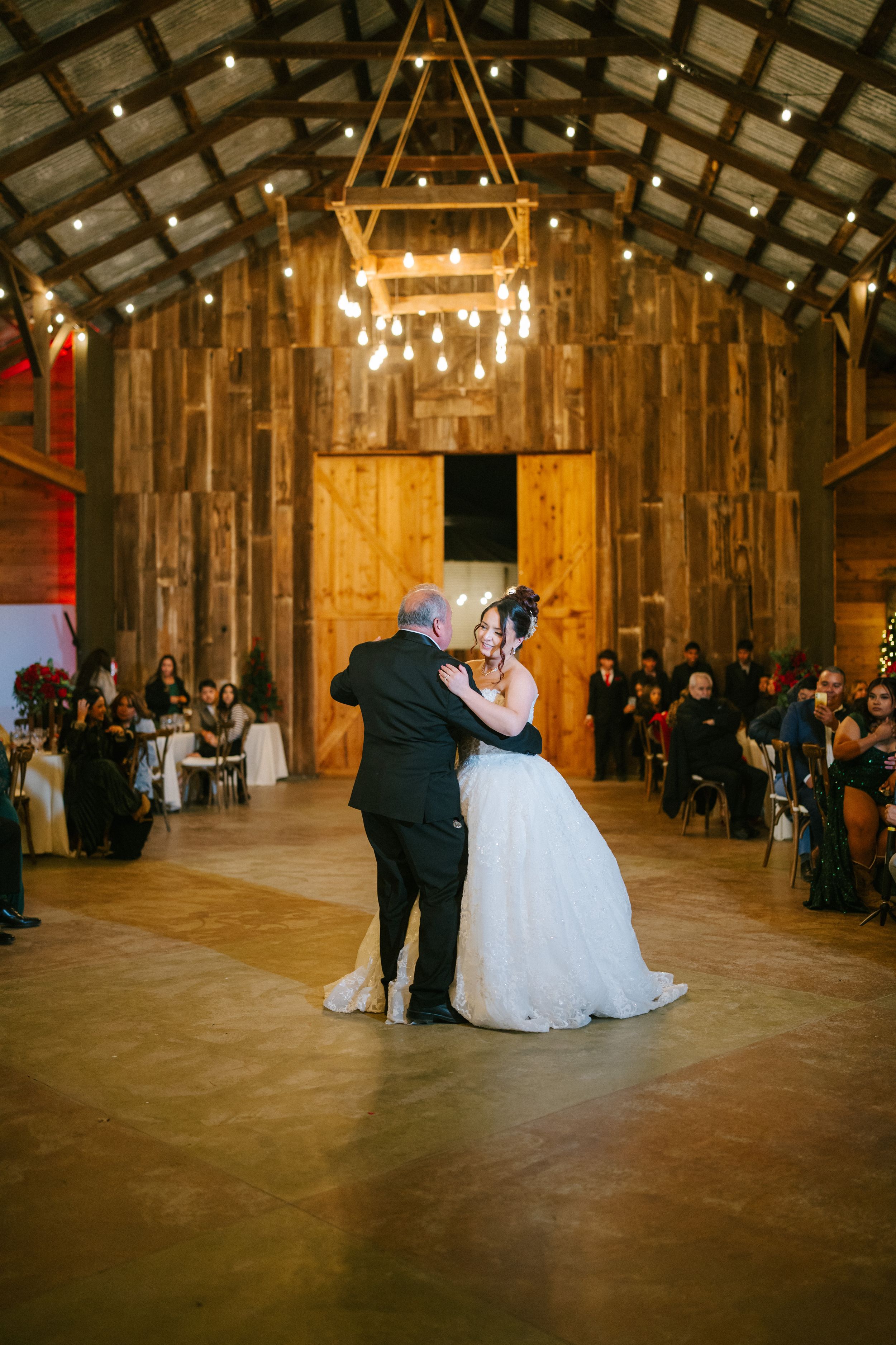 A romantic first dance between newlyweds in a rustic wooden barn venue illuminated by warm overhead lighting.