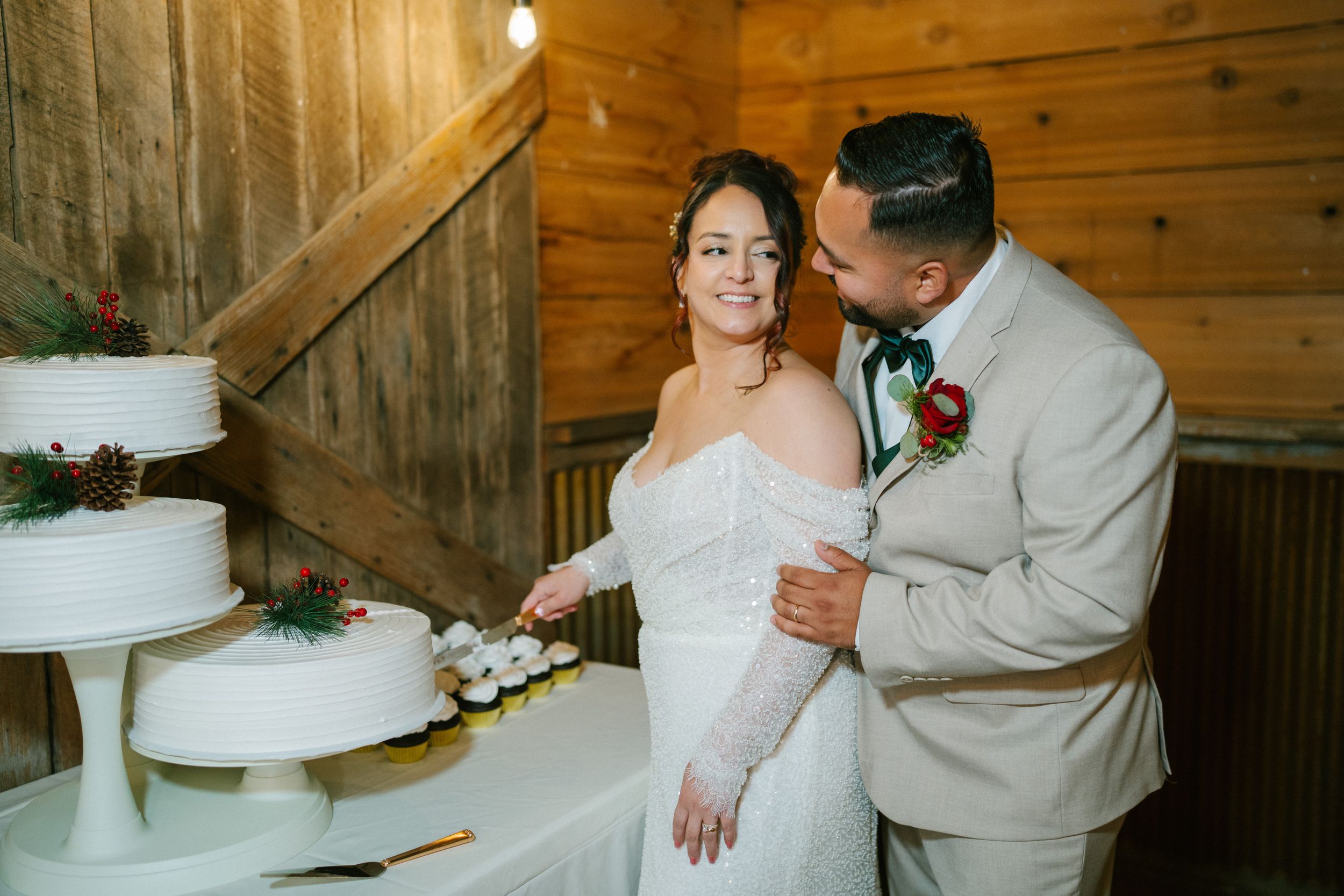 A couple in wedding attire shares an intimate moment beside their white tiered cake in a rustic wooden venue.