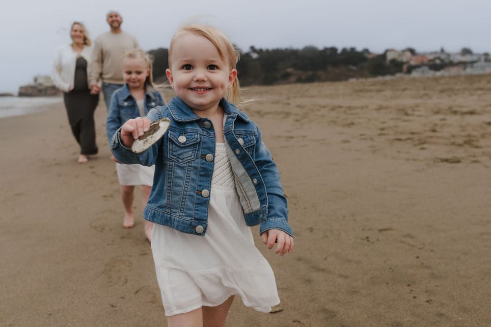 Small child runs joyfully on beach wearing denim jacket and white dress.