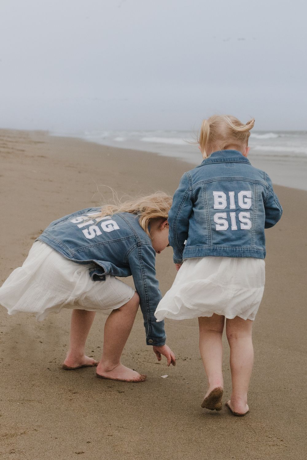Two children wearing matching 'BIG SIS' denim jackets play on beach.