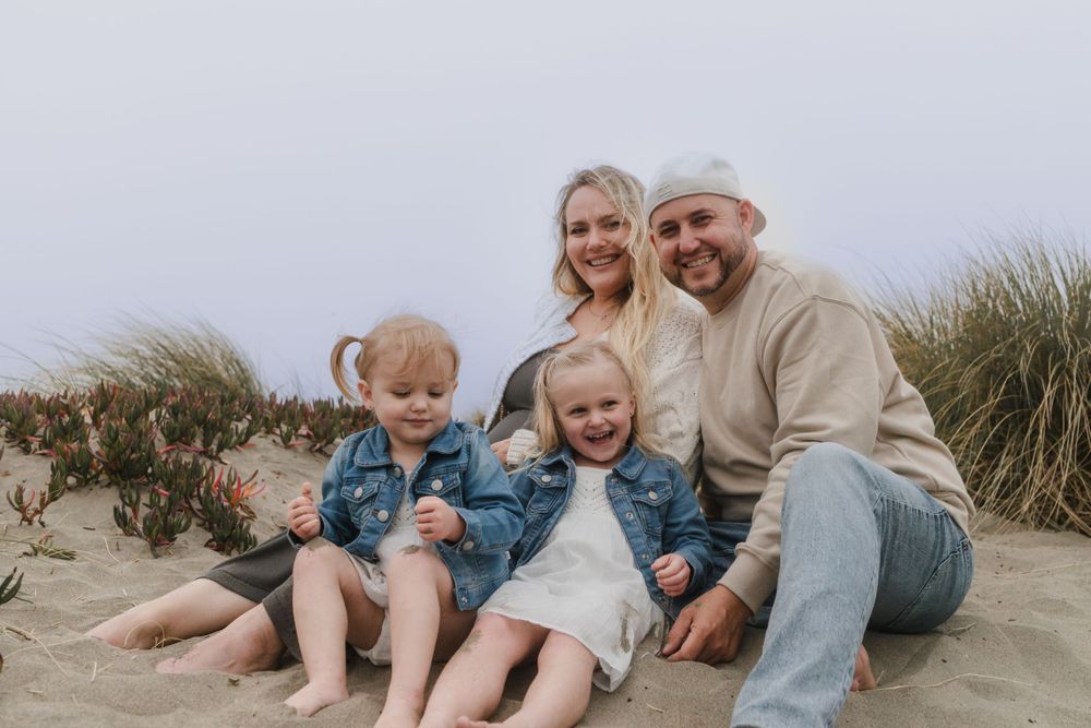 Family sits together on sandy beach dunes surrounded by tall grass.