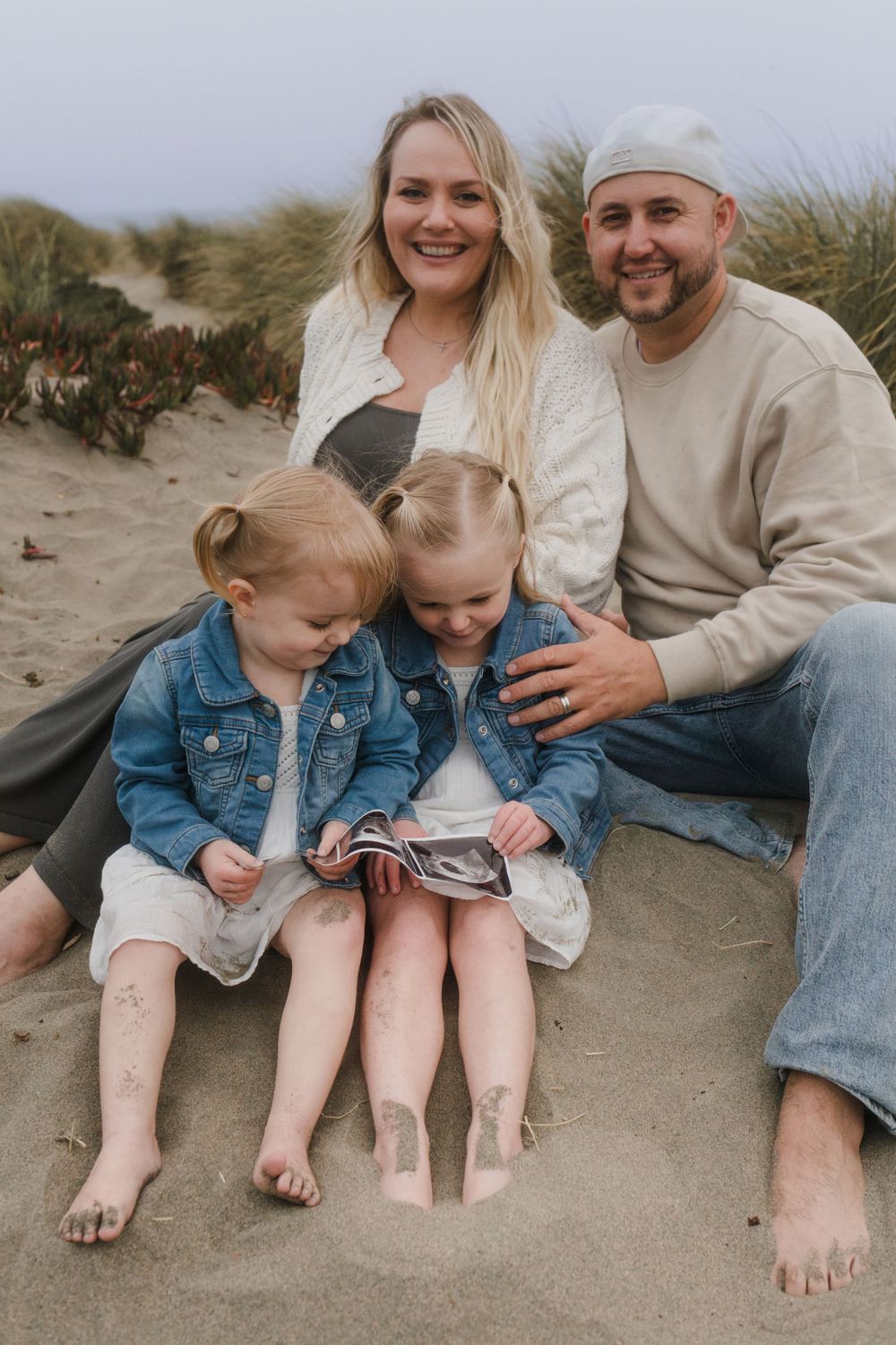 Family sits together on sand dunes while children look at a book.