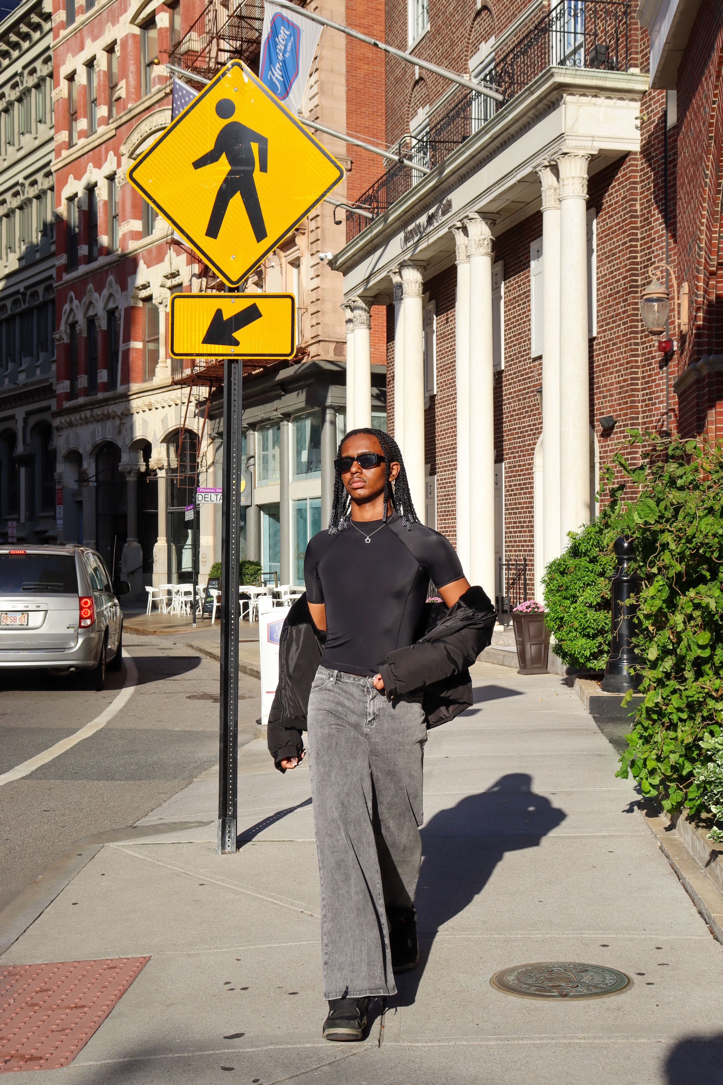 Stylish person stands on city sidewalk beneath yellow pedestrian crossing sign.