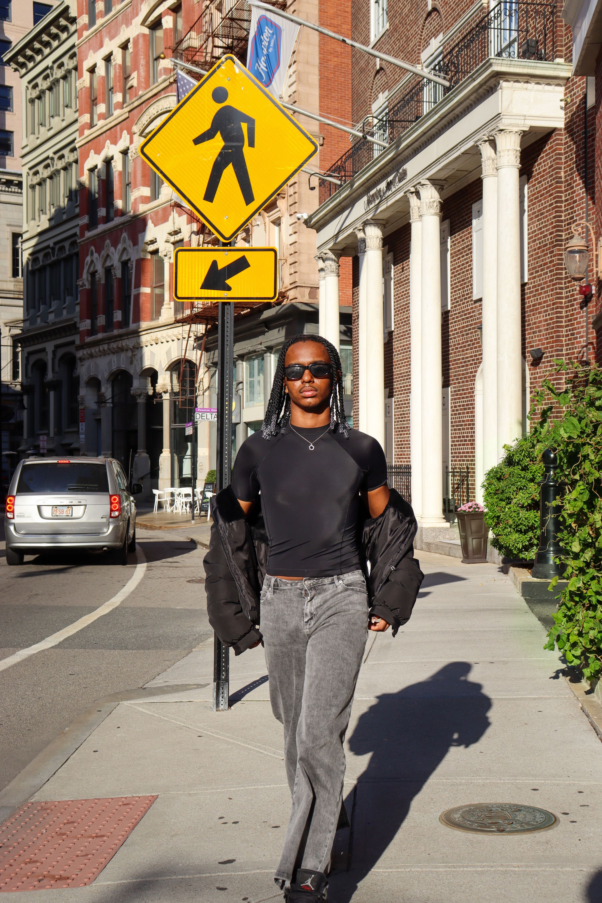 Person poses confidently on city sidewalk near pedestrian crossing sign in urban setting.