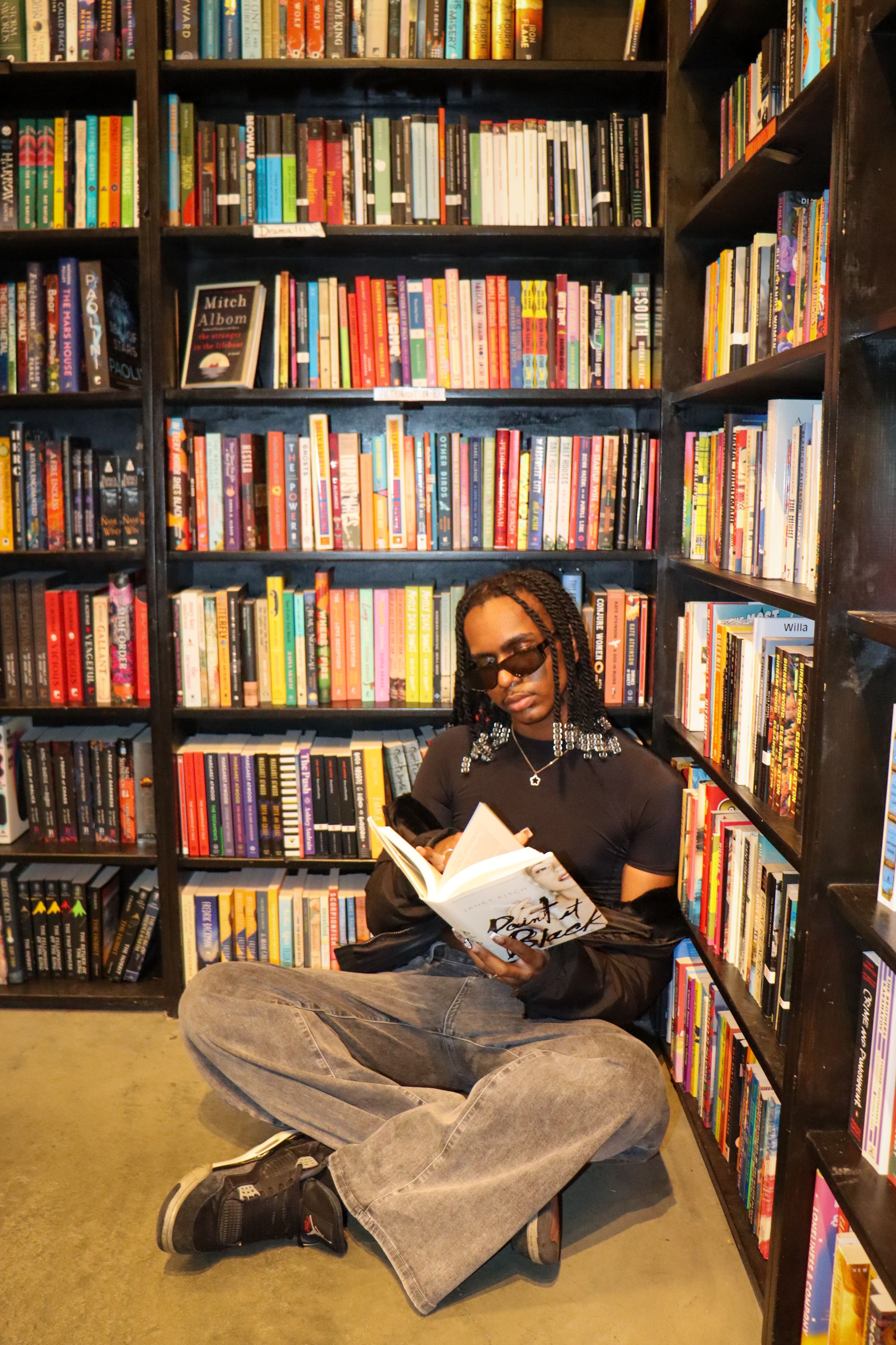Person sits cross-legged reading book surrounded by floor-to-ceiling bookshelves in library.