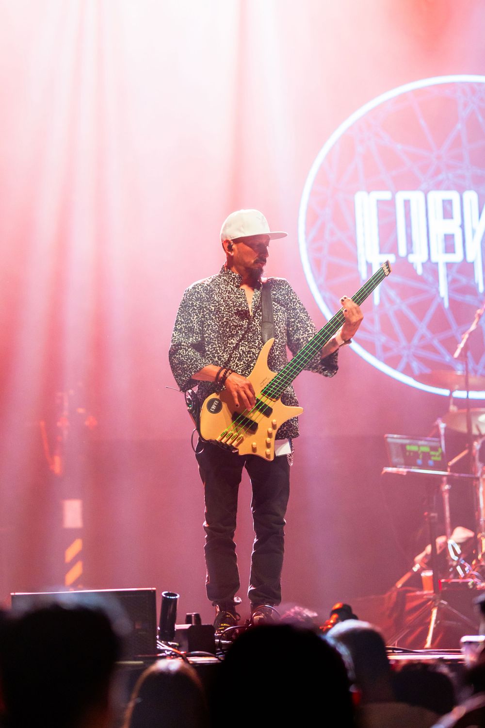 Bass guitarist performs on stage in patterned shirt and cap with illuminated logo behind.