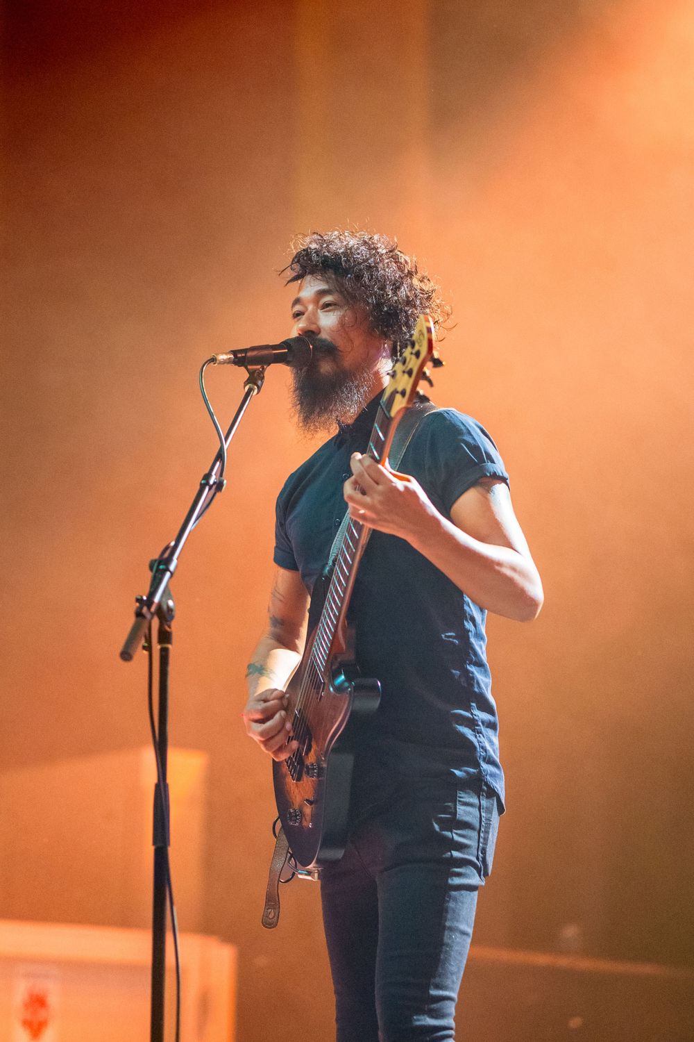 Musician with guitar sings into microphone against orange-lit backdrop.
