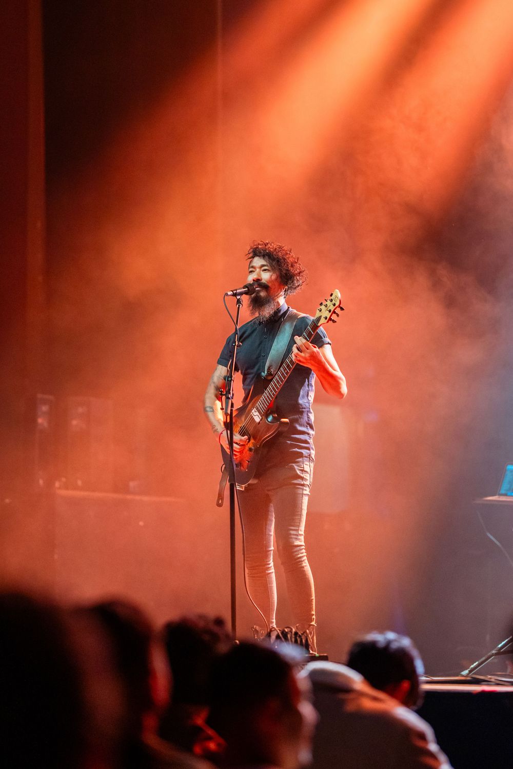 Musician performing on stage with guitar against dramatic orange lighting and silhouetted crowd.