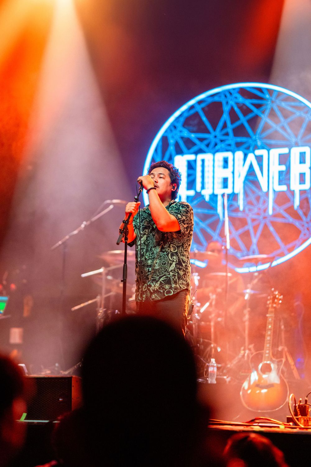 Performer in sparkly green outfit singing on stage with blue geometric logo backdrop and dramatic lighting.