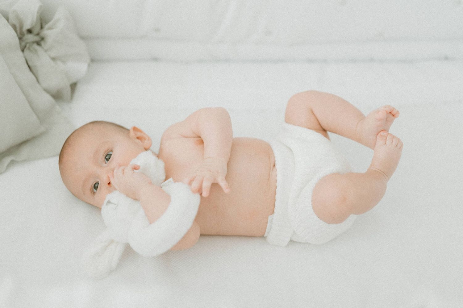 Baby in white diaper lying on back against white bedding.