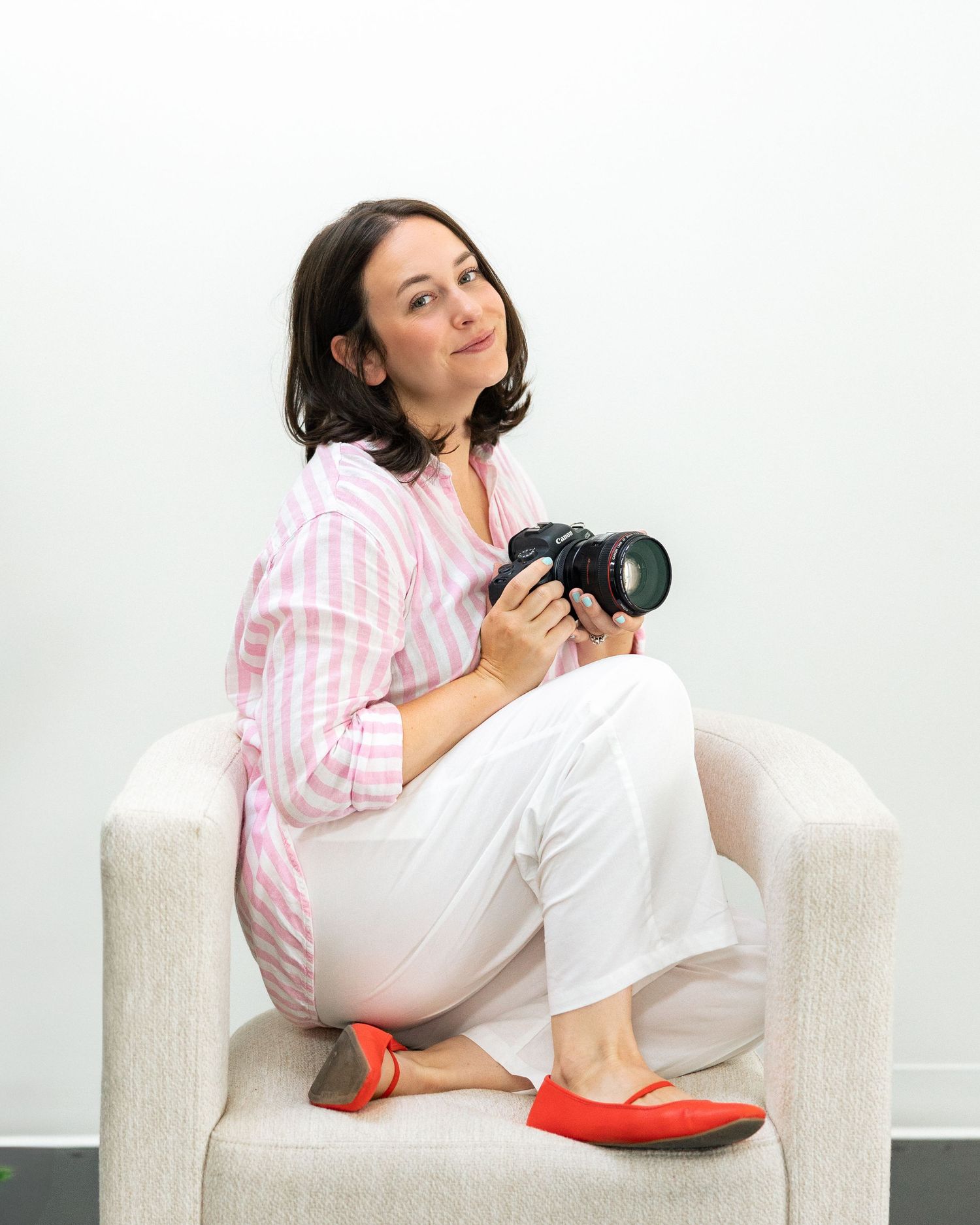 A woman in a pink striped shirt and white pants sits in a chair holding a DSLR camera, looking up thoughtfully.