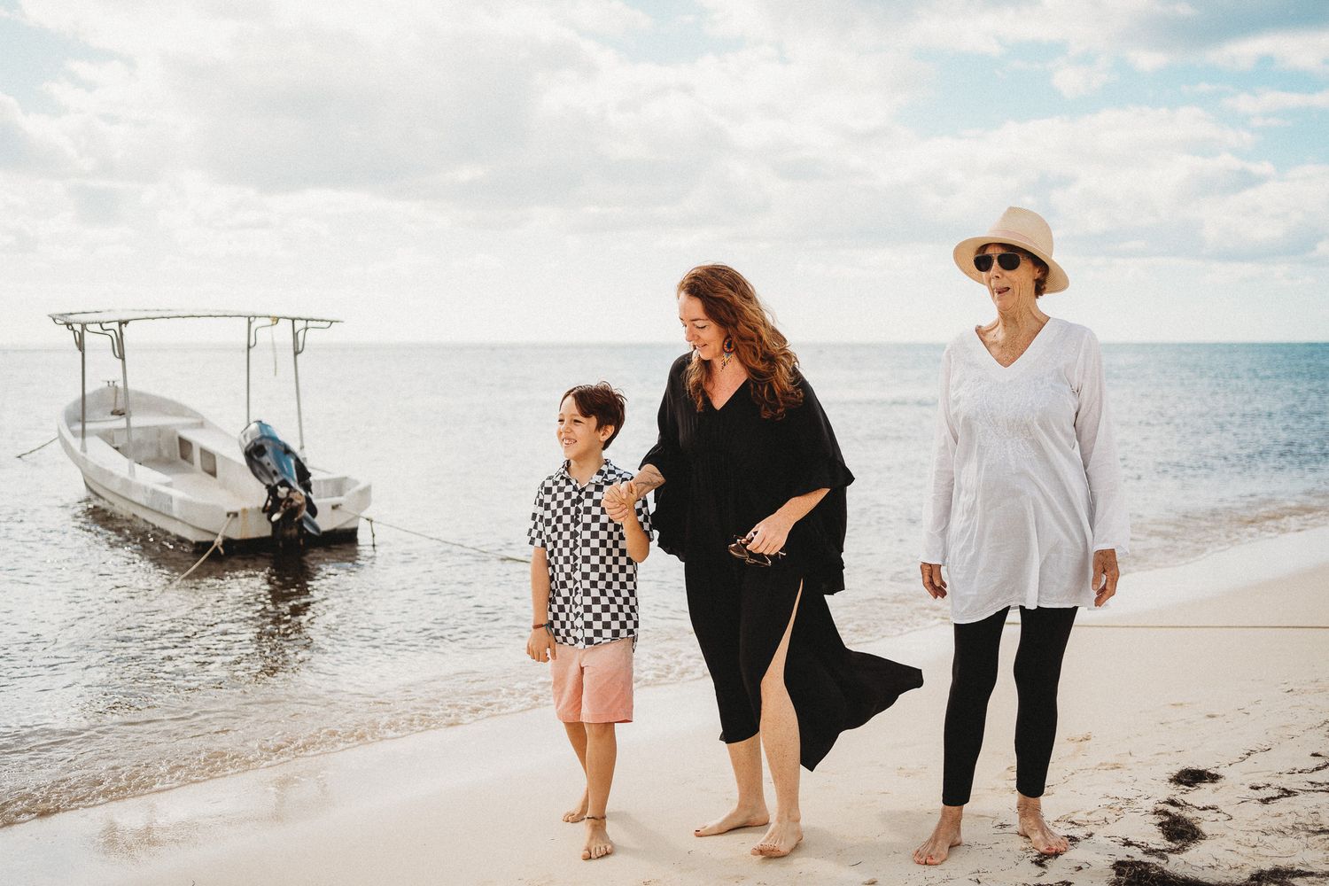Three people stroll along a beach shoreline with a small boat moored in shallow water on a sunny day. Puerto Morelos Family Photography.