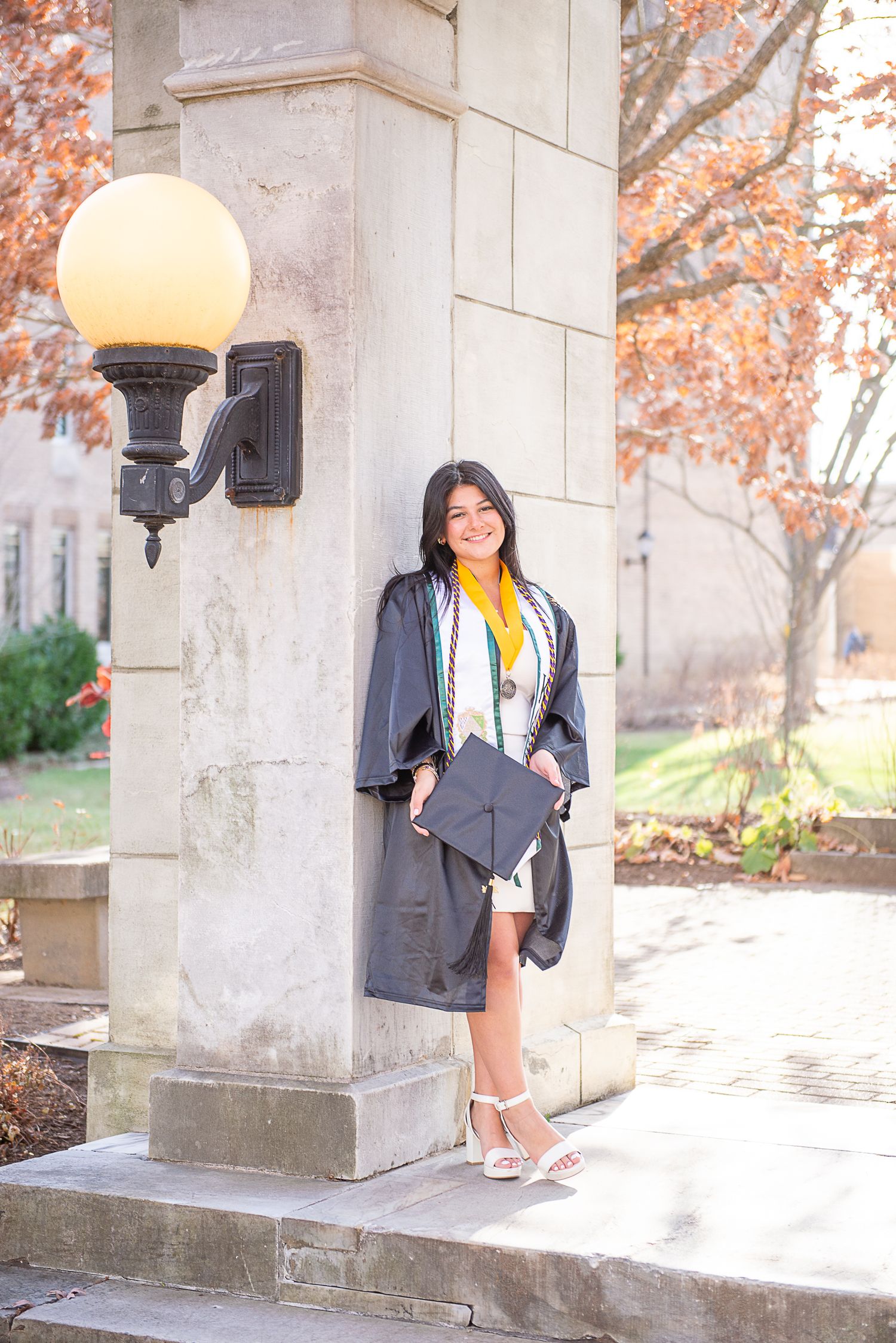 A person in stylish fall fashion poses against stone columns with autumn leaves in the background.