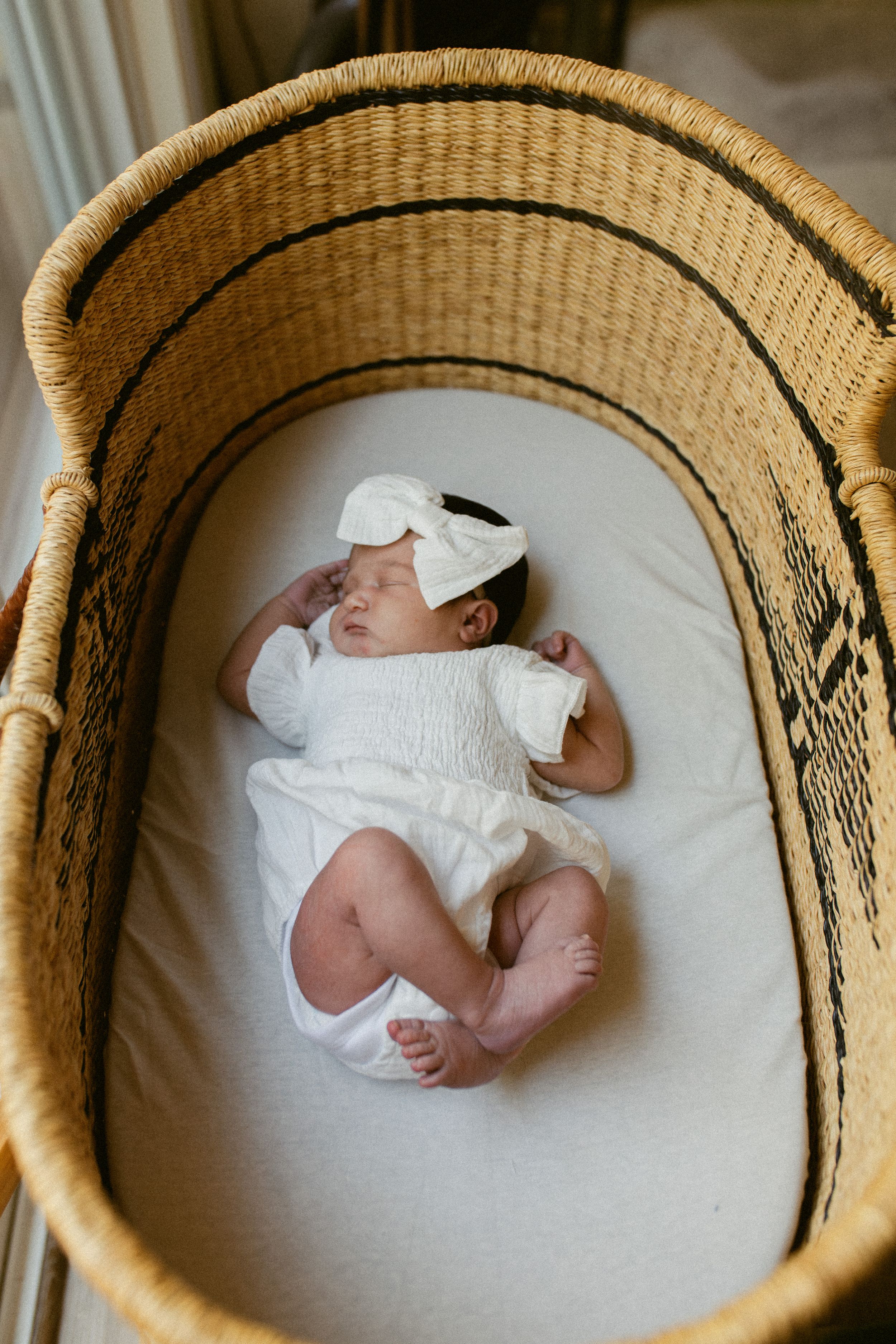 A sleeping newborn wearing white clothing rests peacefully in a woven bassinet with beige bedding.