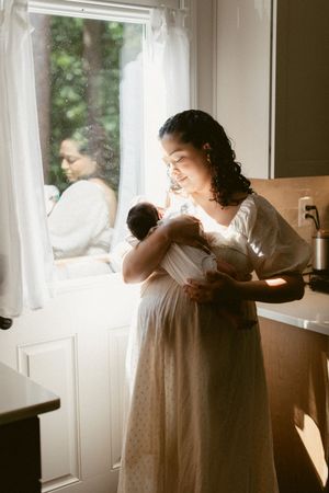 Soft sunlight streams through window as figure in flowing white dress holds and nurtures infant in peaceful home setting.
