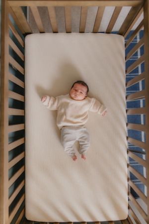 Overhead view of infant wearing cream colored outfit lying in wooden crib.