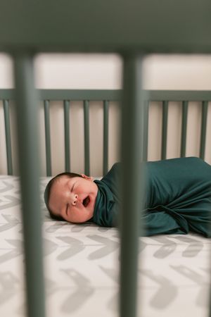 A sleeping baby wearing a dark green sleeper lies in a white crib with chevron patterned bedding.