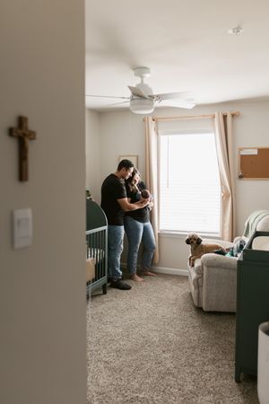 A cozy nursery room features a ceiling fan, carpeted floor, cross on wall, and a dog watching from the corner.