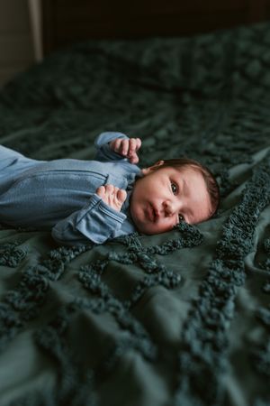 A baby in a blue outfit lies on a textured dark green bedspread while looking at the camera.