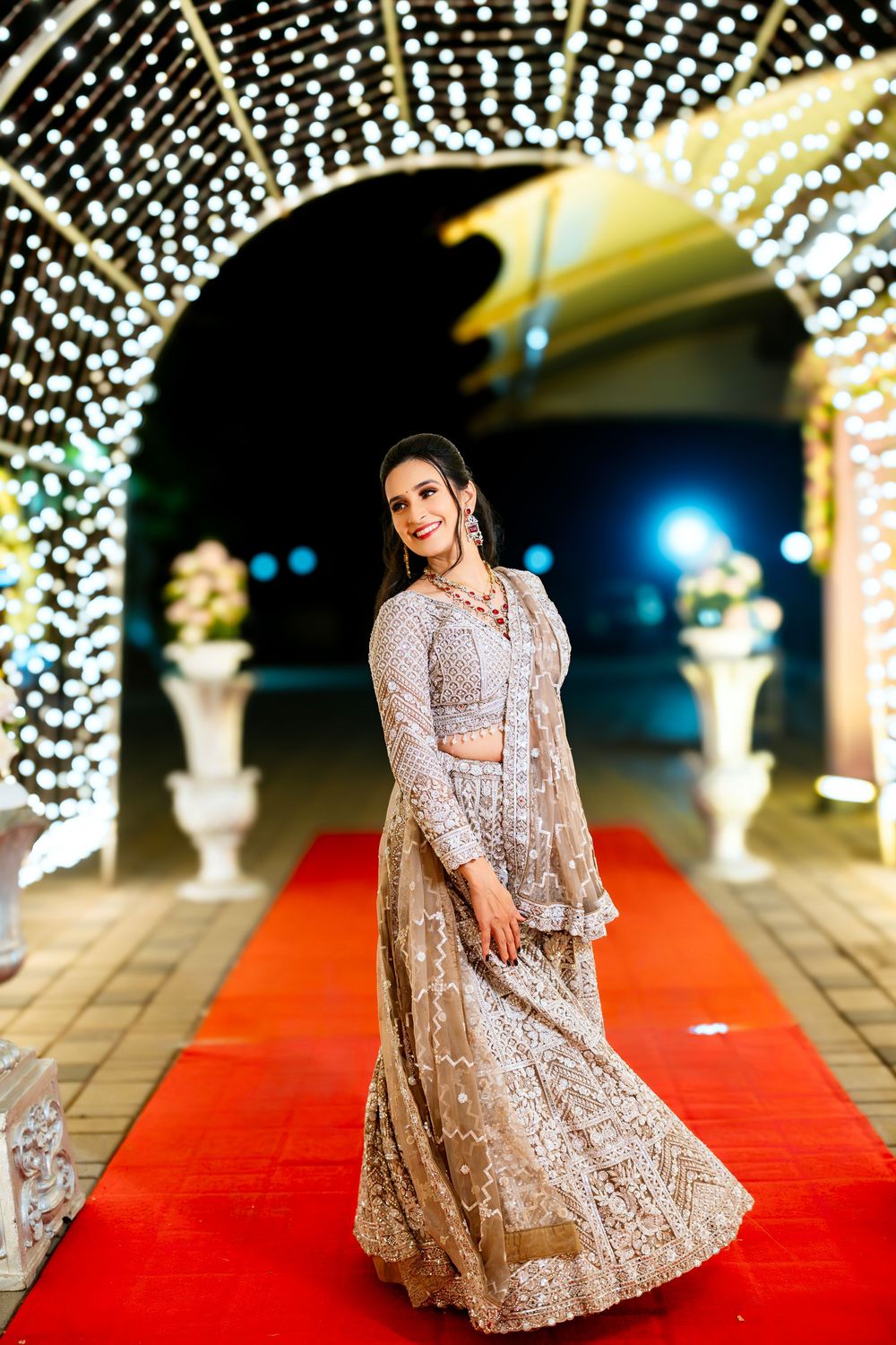 Bride in sparkly gown poses elegantly on red carpet under canopy of string lights.
