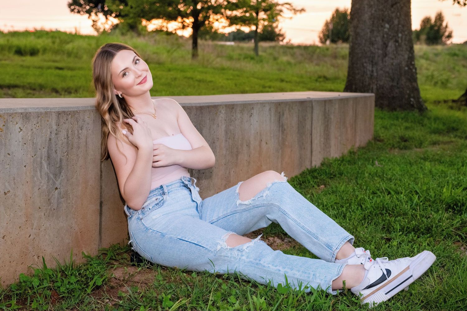 Someone in ripped jeans and a pink top relaxes on a concrete ledge at sunset in a grassy park area.