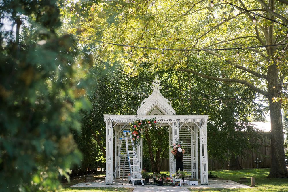 A white garden gazebo surrounded by lush greenery creates a romantic outdoor setting in filtered sunlight.