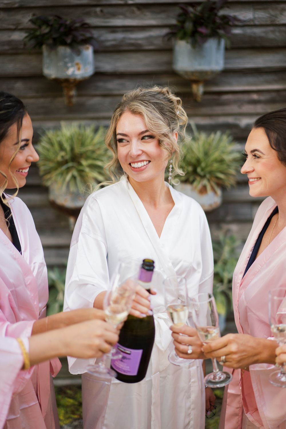 Wedding party members in white and pink robes smile and share drinks together during pre-ceremony preparations.