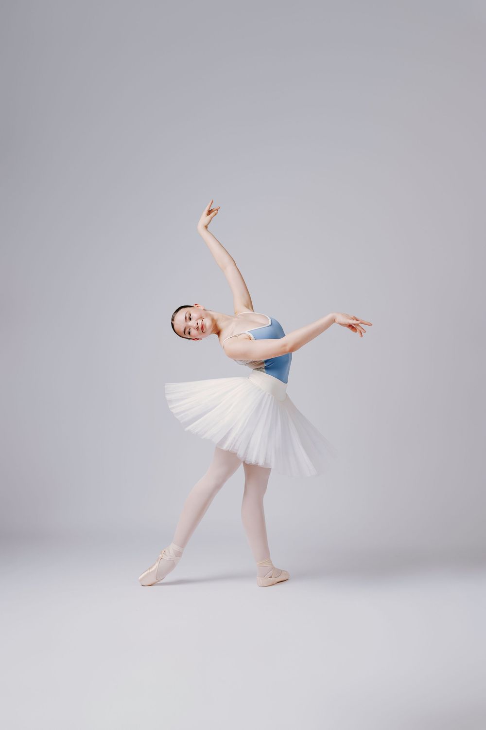 A ballet dancer in a white tutu and blue leotard performs graceful poses in a minimalist studio setting.