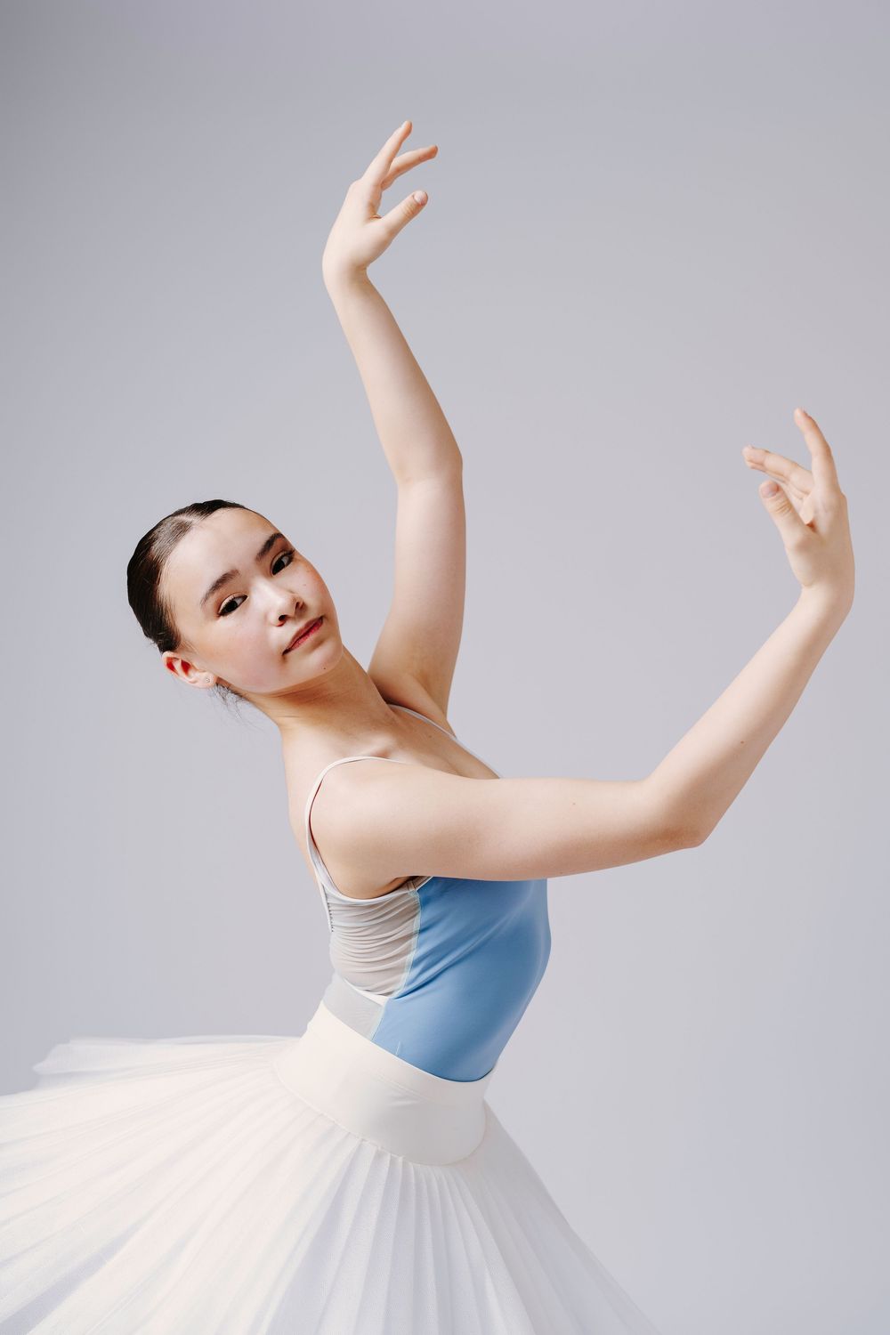 Classical ballet dancer poses in blue and white costume with elegant arm positions against neutral background.