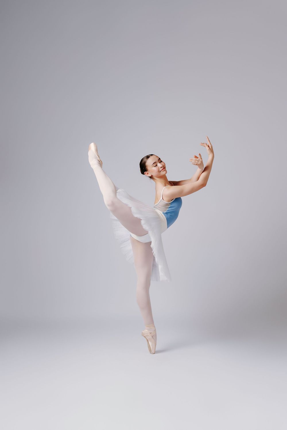 Ballet dancer executing an elegant arabesque position in blue leotard and white tulle skirt.