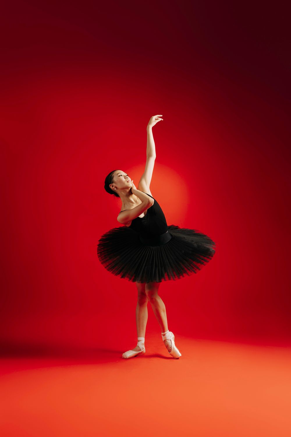 Ballerina in black tutu performs elegant poses against dramatic red studio background.