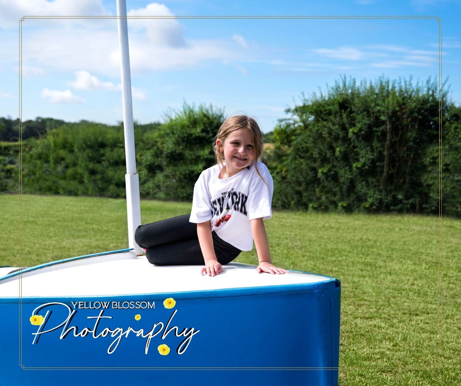 A youth athlete in a white jersey sits by a soccer goal post on a sunny day at a sports field.
