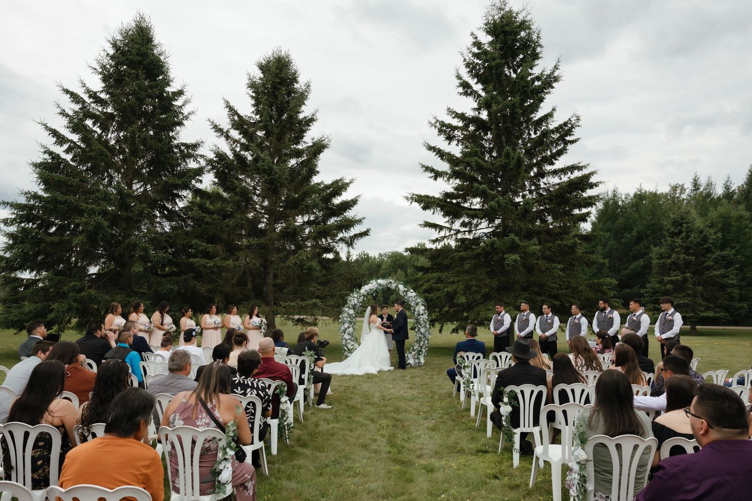 A large joyful wedding party celebrates and laughs together during an indoor reception at a rustic barn-style venue.