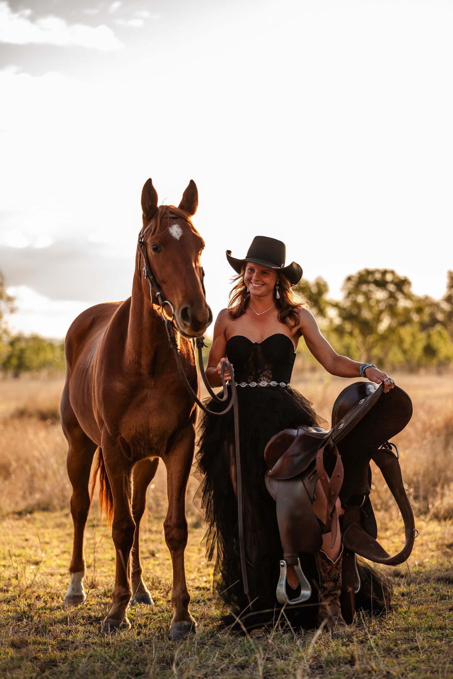 A dog and horse pose together in a golden field during sunset while their owner stands nearby in western attire.