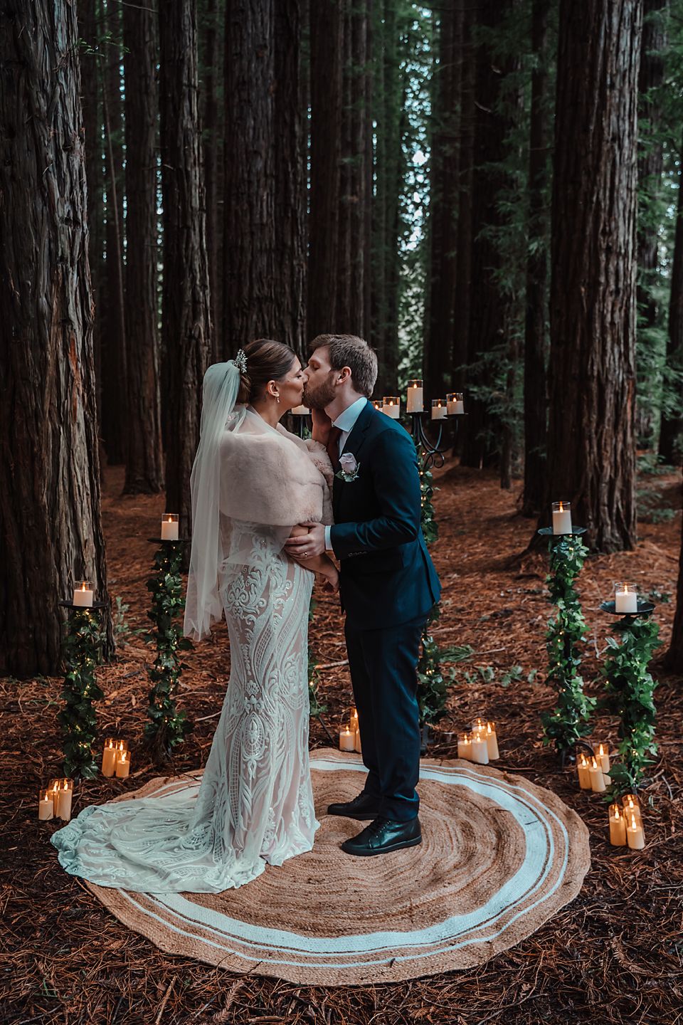 A romantic forest wedding ceremony with a decorative rug path surrounded by glowing lanterns and towering redwood trees.