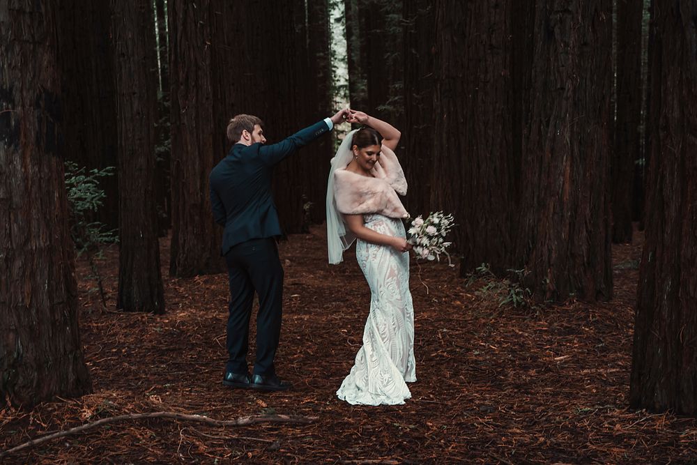 Wedding couple walks through a redwood forest surrounded by towering trees and fallen leaves.